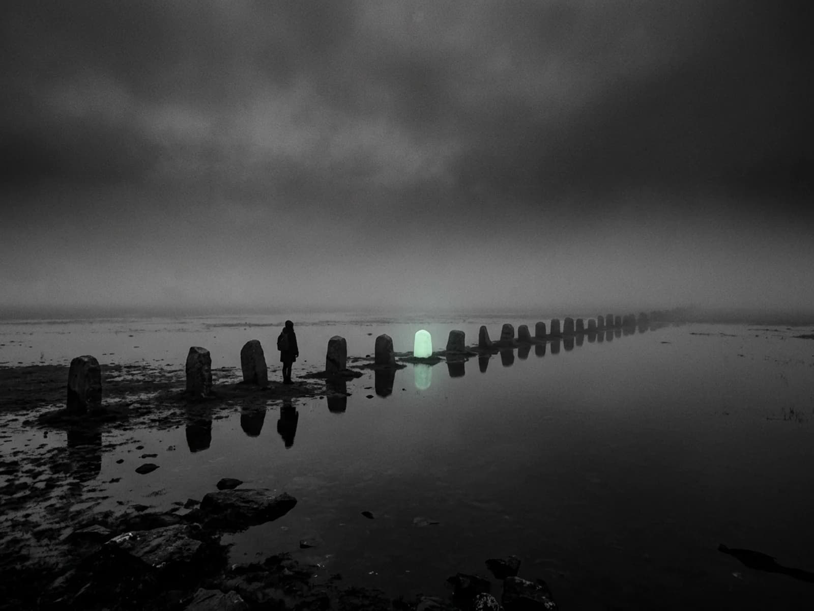 A chain of stone markers crosses a dark flooded plain at night with one marker glowing pale mint, and a small figure standing beside it